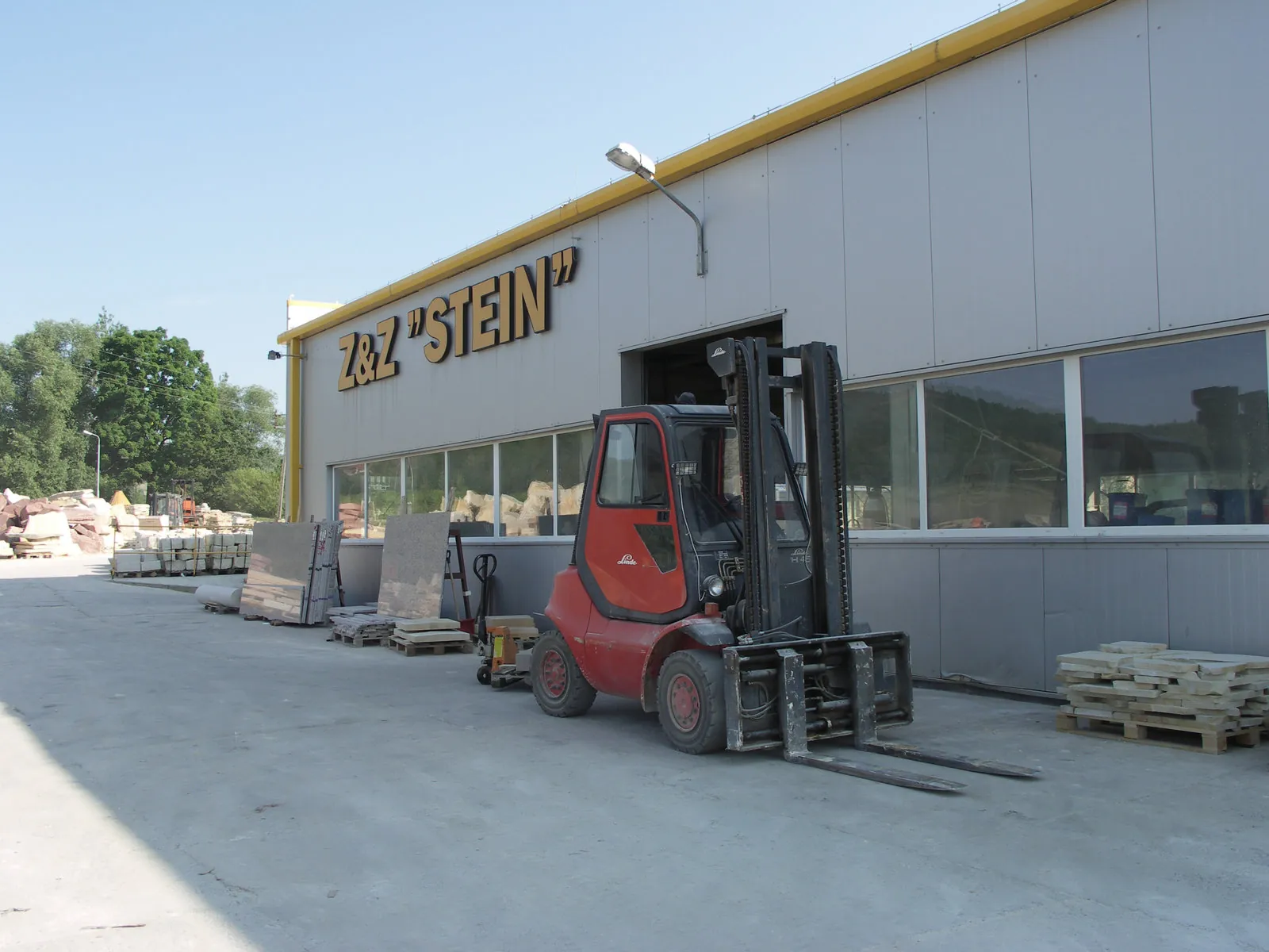 Produktionshalle Z&Z Stein mit Gabelstapler im Betrieb, Sandstein-Paletten und Rohmateriallager auf dem Betriebsgelände – Außenansicht Werkstattgebäude